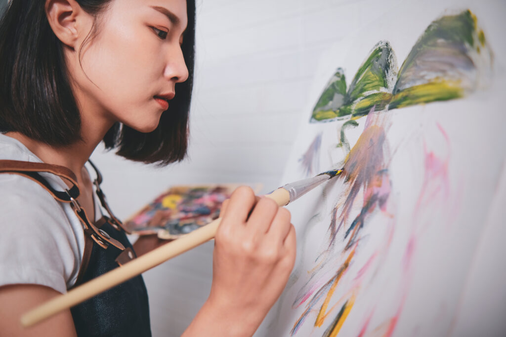 Artist Painting Picture With Blank Canvas Side View, A young woman with short dark hair is painting on a canvas. She holds a palette in her left hand and a brush in her right, creating a colorful landscape with green and yellow mountains. She wears a dark apron and focuses intently on her work.