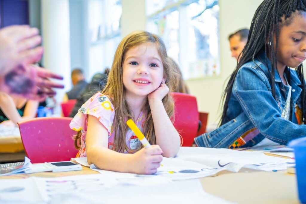 little girl smiles at the camera, proud of the artwork she just created in one of the High's workshops.