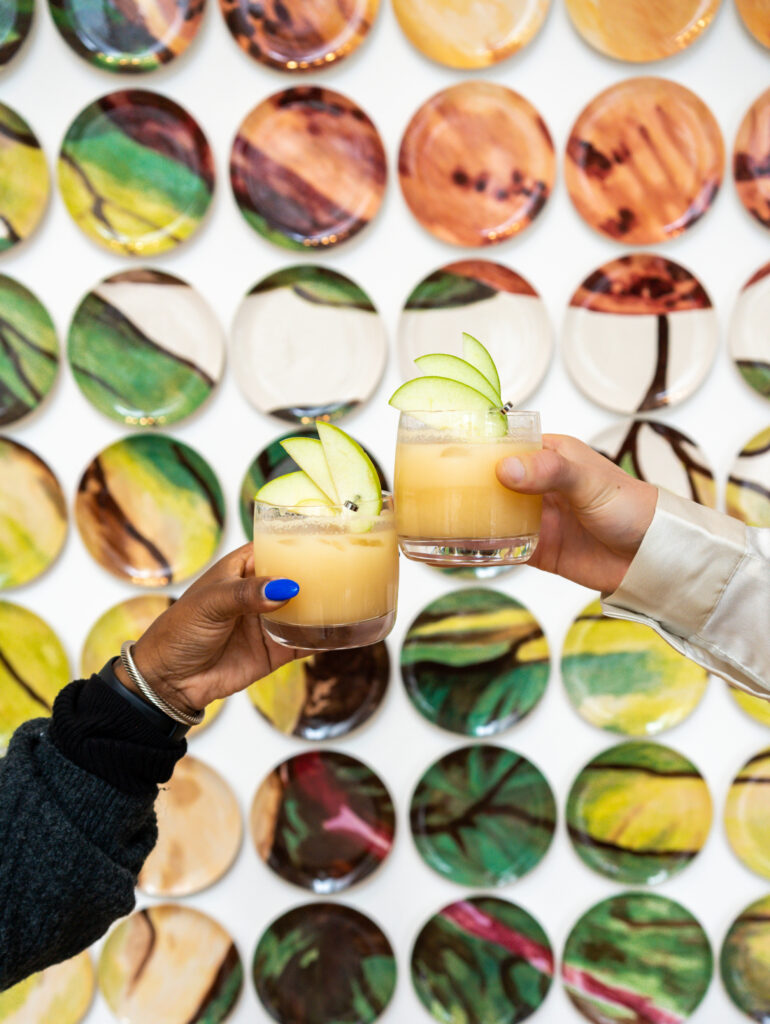 Two hands cheers an apple cocktail together with a wall of decorative plates behind them