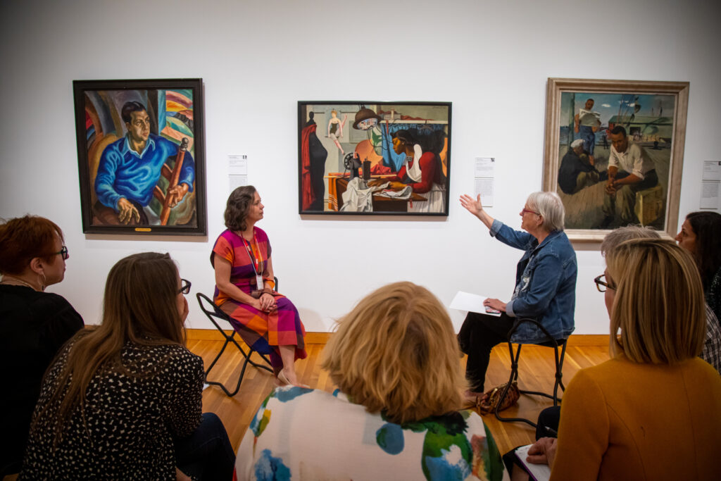 Eve Caa 2023186 O2 A group of women sit on stools while looking toward a painting of a woman sewing
