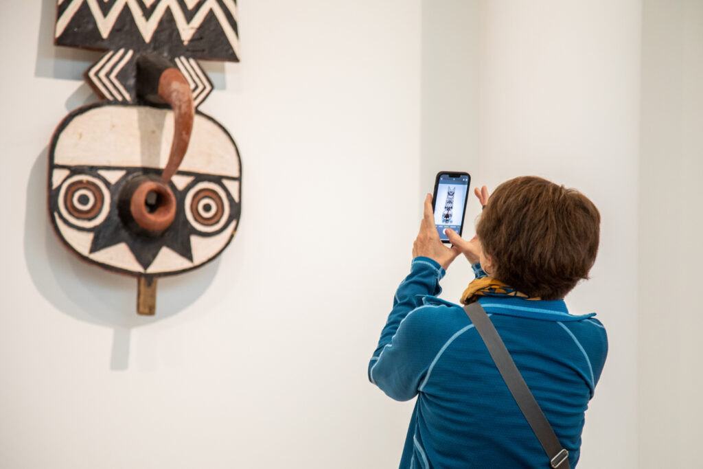 A person in a blue jacket is taking a photo of a tribal mask on display using a smartphone. The mask features geometric patterns and bold colors, with a prominent curved element at the bottom. The setting appears to be a museum or gallery.