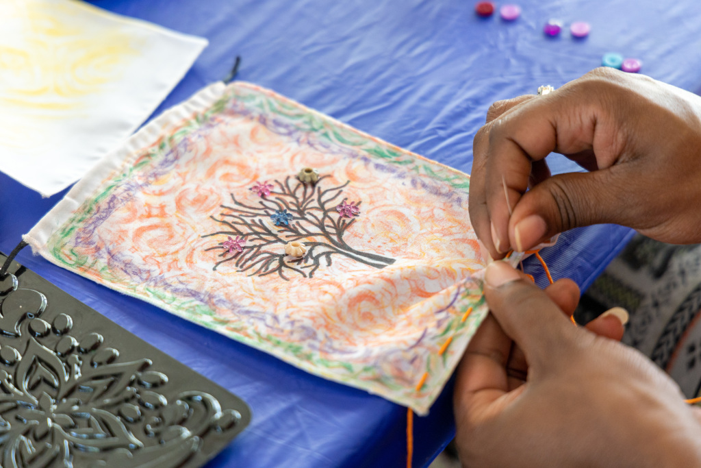 A person is sewing on a piece of cloth decorated with a tree and colorful designs. Their hands are visible as they stitch, surrounded by a blue tablecloth and art supplies such as buttons and a stencil.