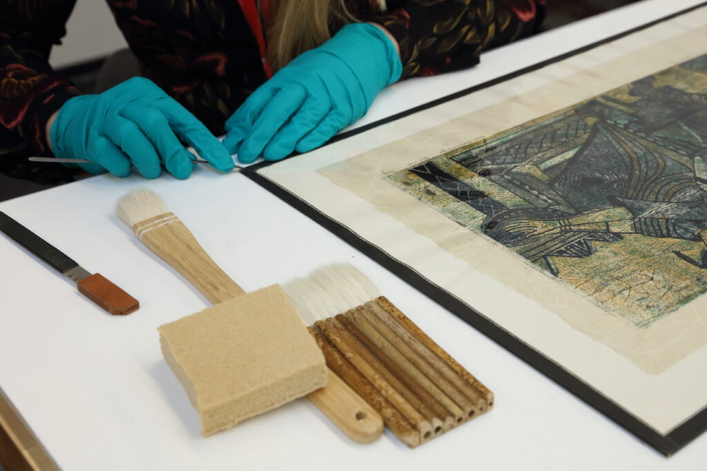 A conservator wears gloves and touches an artwork while a sponge and soft bristle brush are laid on the table
