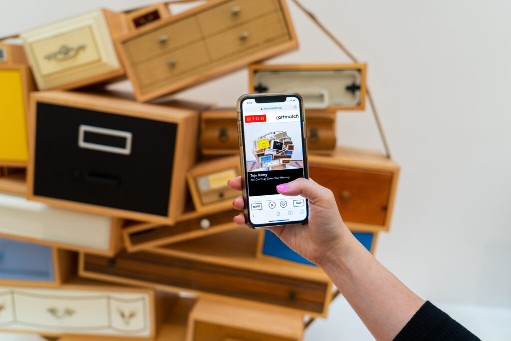 a woman holds a phone displaying the web app, heartmatch, in front of a cabinet by Tejo Remy.