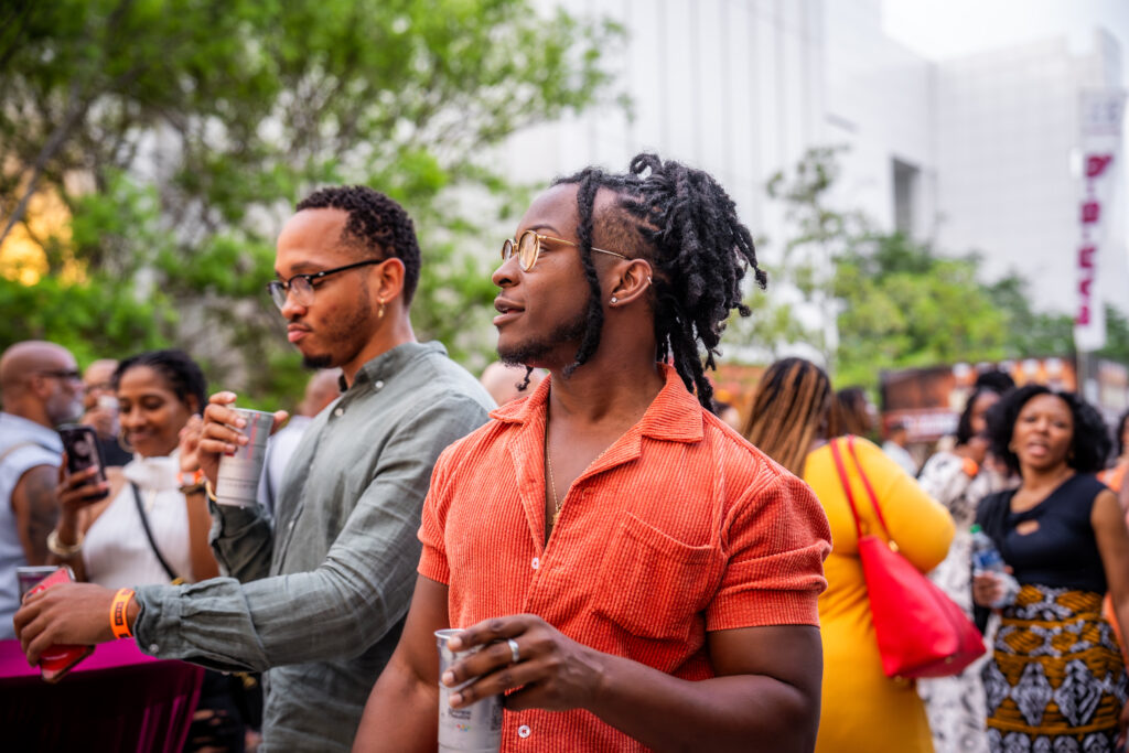 A lively outdoor gathering with people socializing. A man in an orange shirt holds a cup, looking to the side. Another person in glasses stands nearby. The background features trees and a building, with several attendees visible.