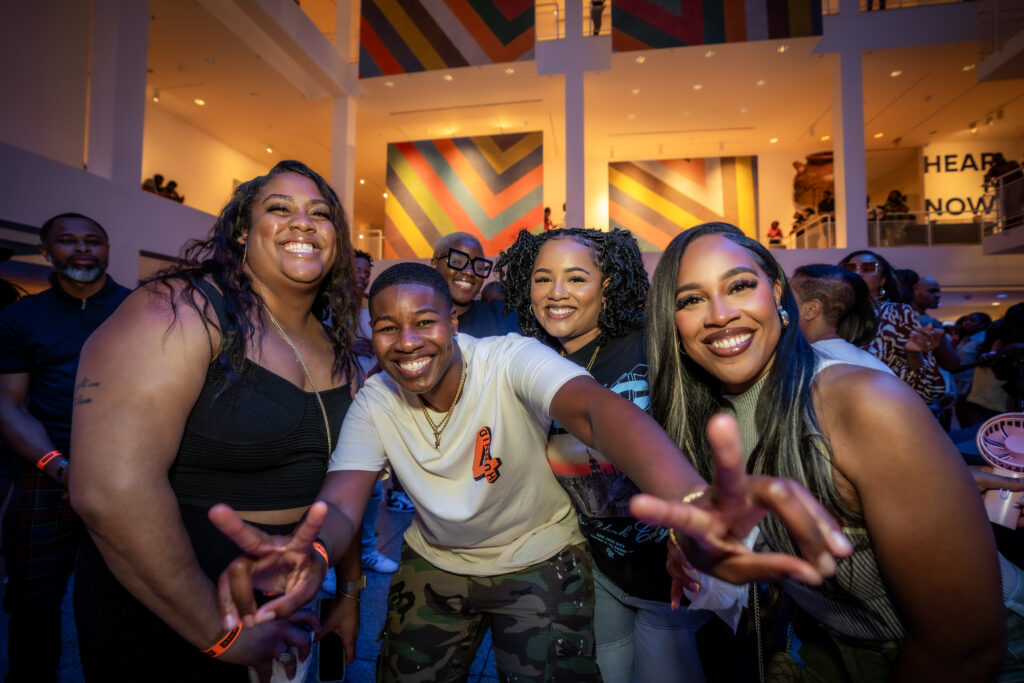 A group of people smiling and posing for a photo at an indoor event, with colorful abstract art displayed on the walls behind them. The atmosphere is lively and cheerful.