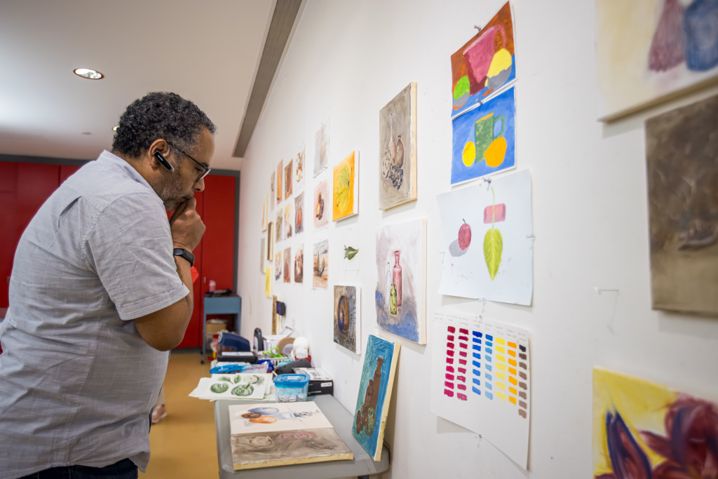 A man with glasses and a headset observes a wall displaying colorful artwork, including fruits and bottles. The art pieces are various sizes, some with color swatches. The room has red walls and a table with art supplies.