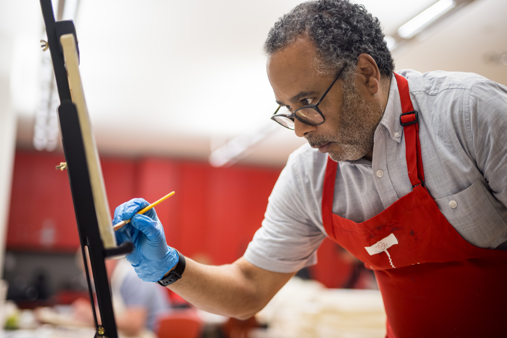 A person wearing a red apron and blue gloves is focused on painting on a canvas, using a detail brush in a well-lit room.