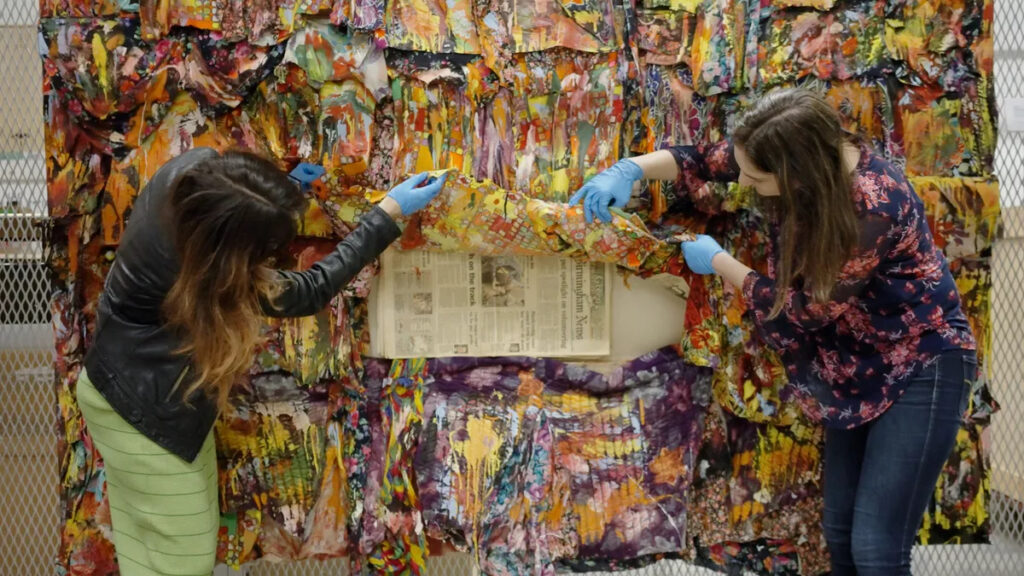 Two women with blue gloves inspect a colorful work of folk art made from strips of fabric and splattered with paint.