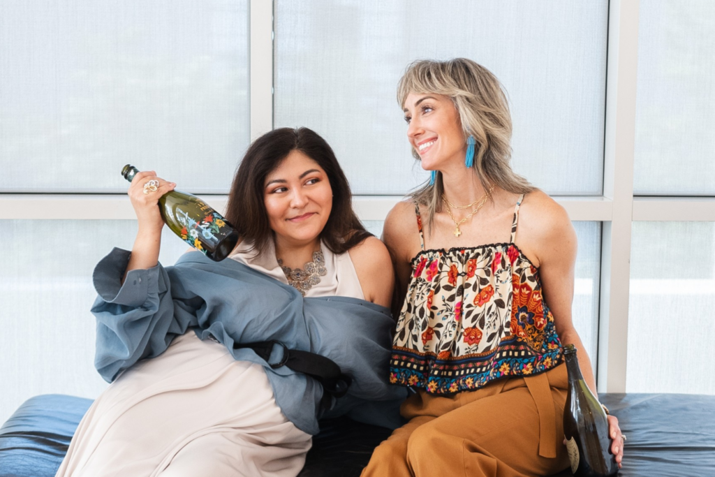 Two women are sitting on a black bench in a bright room. One wears a long white dress and blue shawl, holding a bottle. The other wears a patterned top and brown pants, smiling and looking at her friend. Both have colorful high heels.