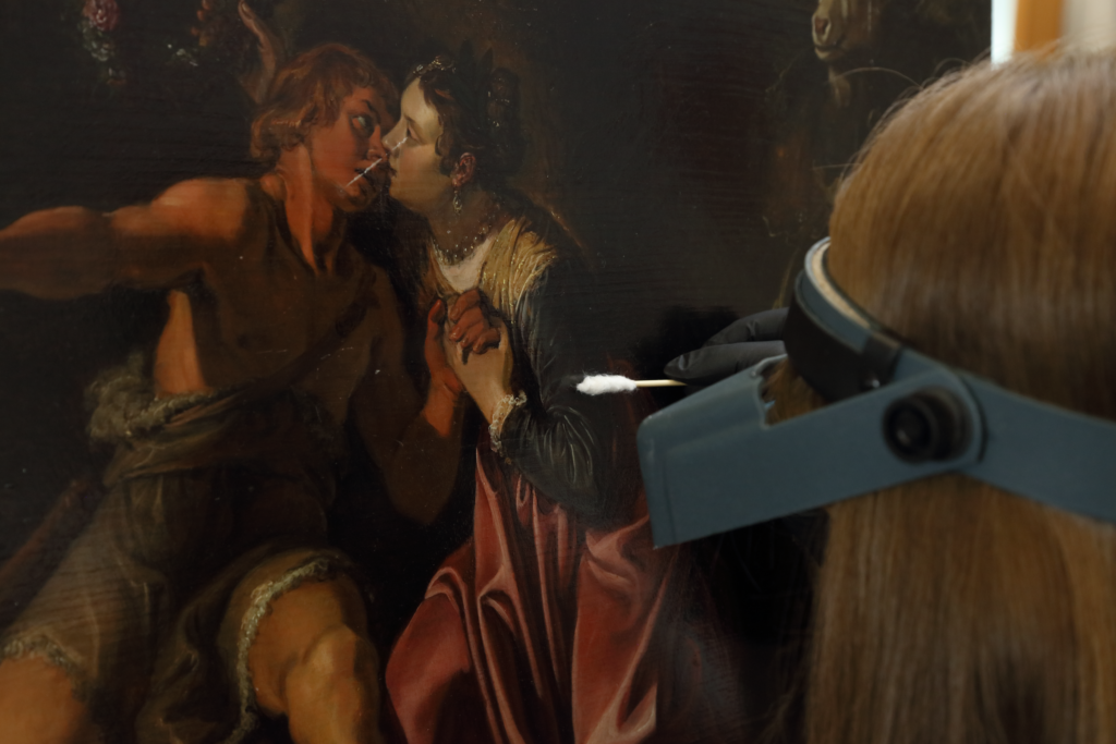 A blonde woman wears a headband magnifier and touches a cotton swab q-tip to a painting
