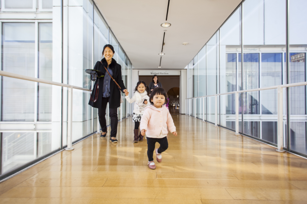 Screenshot 20220707 141656 Young child walking in front of an adult holding a hand of another child in a well lit hallway.
